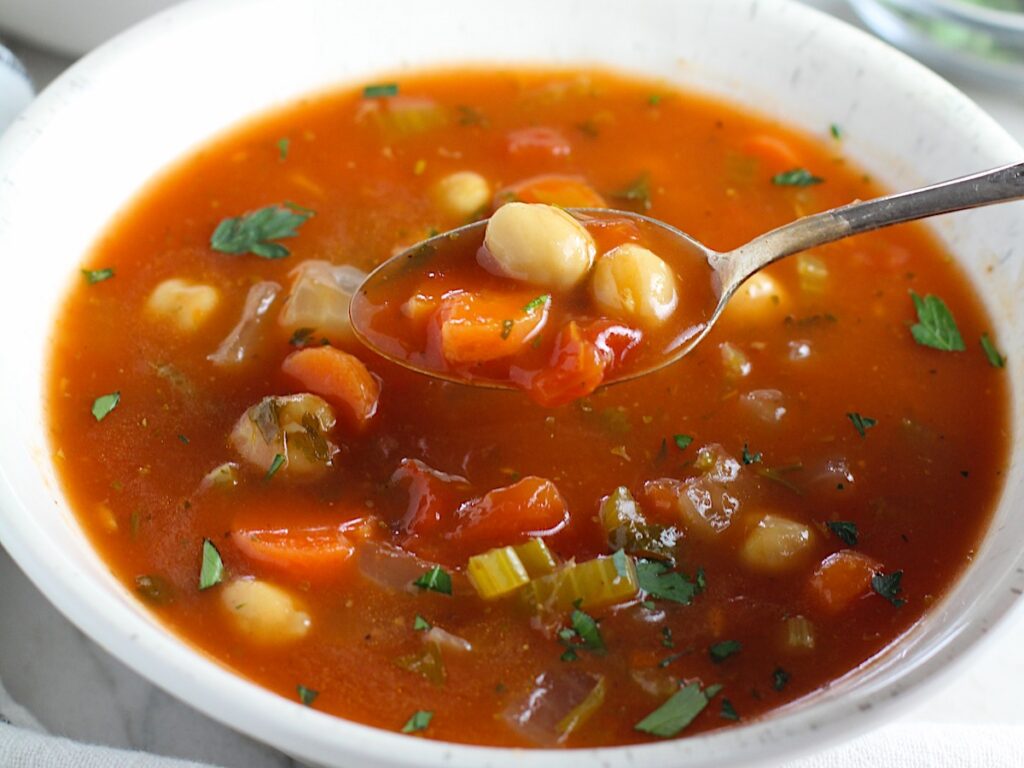 Vegetable Garbanzo Soup recipe in a bowl with spoon filled with soup above. It's slightly creamy and filled with flavor and texture! It's super easy to make ahead and enjoy during the week.