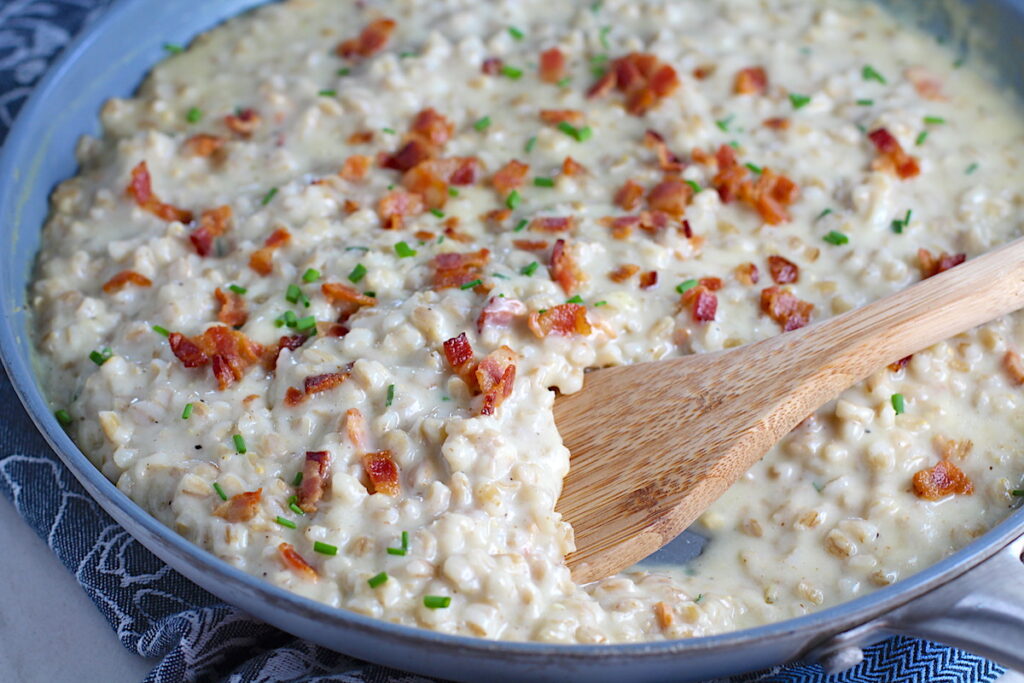 Velvety Barley Recipe with Bacon and Gruyere Cheese in pan with wood spoon and crispy bacon bits and diced chives on top. It's creamy, rich, nutty, smokey, hearty, and utterly delicious.