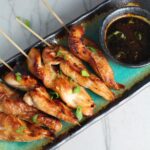 Chinese Chicken on a Stick Recipe on a rectangle platter with small bowl of dipping sauce and pan in background.