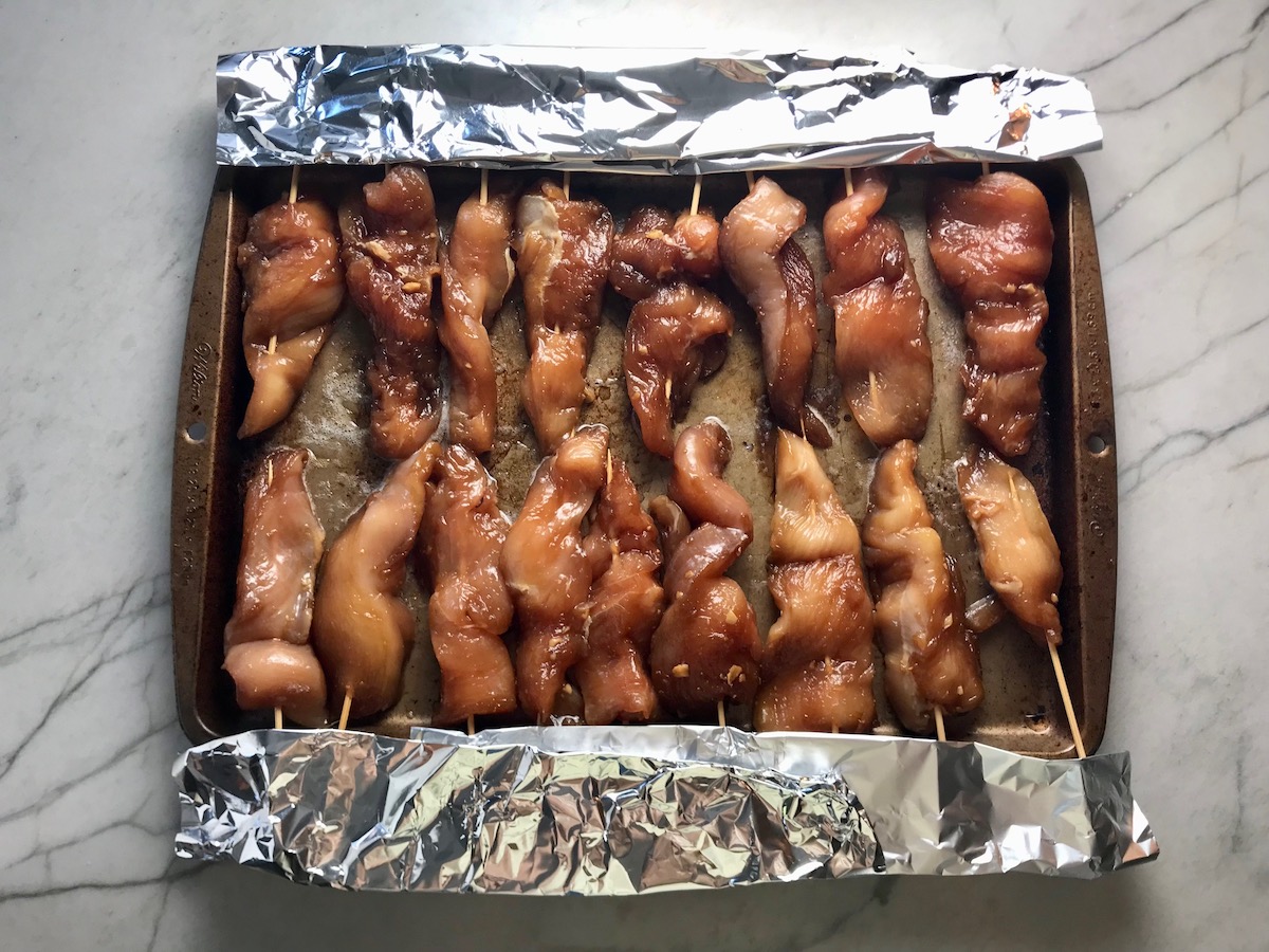 Raw chicken on skewers on a sheet pan with aluminum foil covering the ends of the skewers for Chinese Chicken on a Stick Recipe.