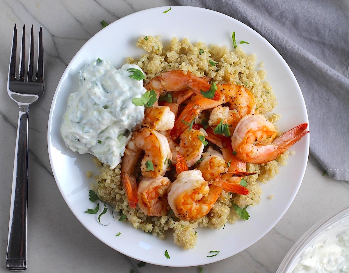 Garlic Lime Grilled Greek Shrimp with Tzatziki Sauce over Quinoa on plate. The Shrimp is simply sauteed with minced garlic, olive oil, and lime for big bold flavors. The Tzatziki Sauce is bright and cool with creamy yogurt with fresh cucumber, lime, garlic, and scallion.