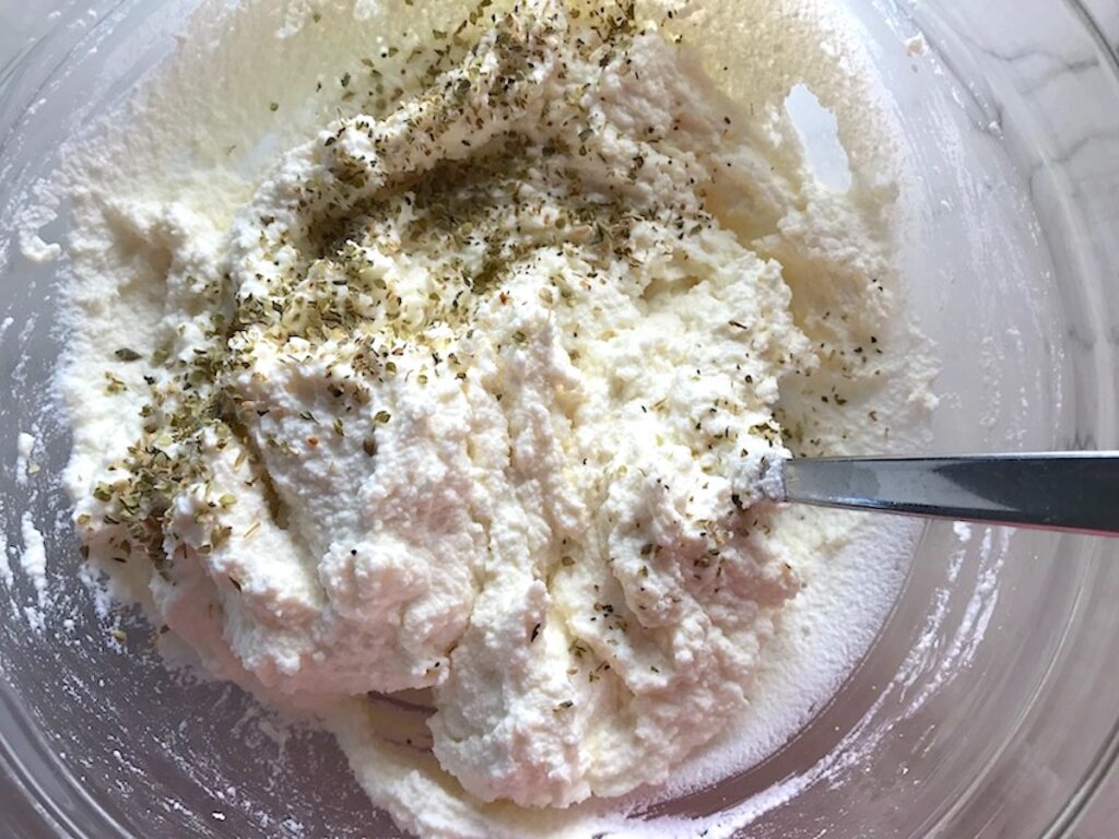 Ricotta Filling recipe being mixed in a clear bowl with italian seasoning and parmesan cheese.