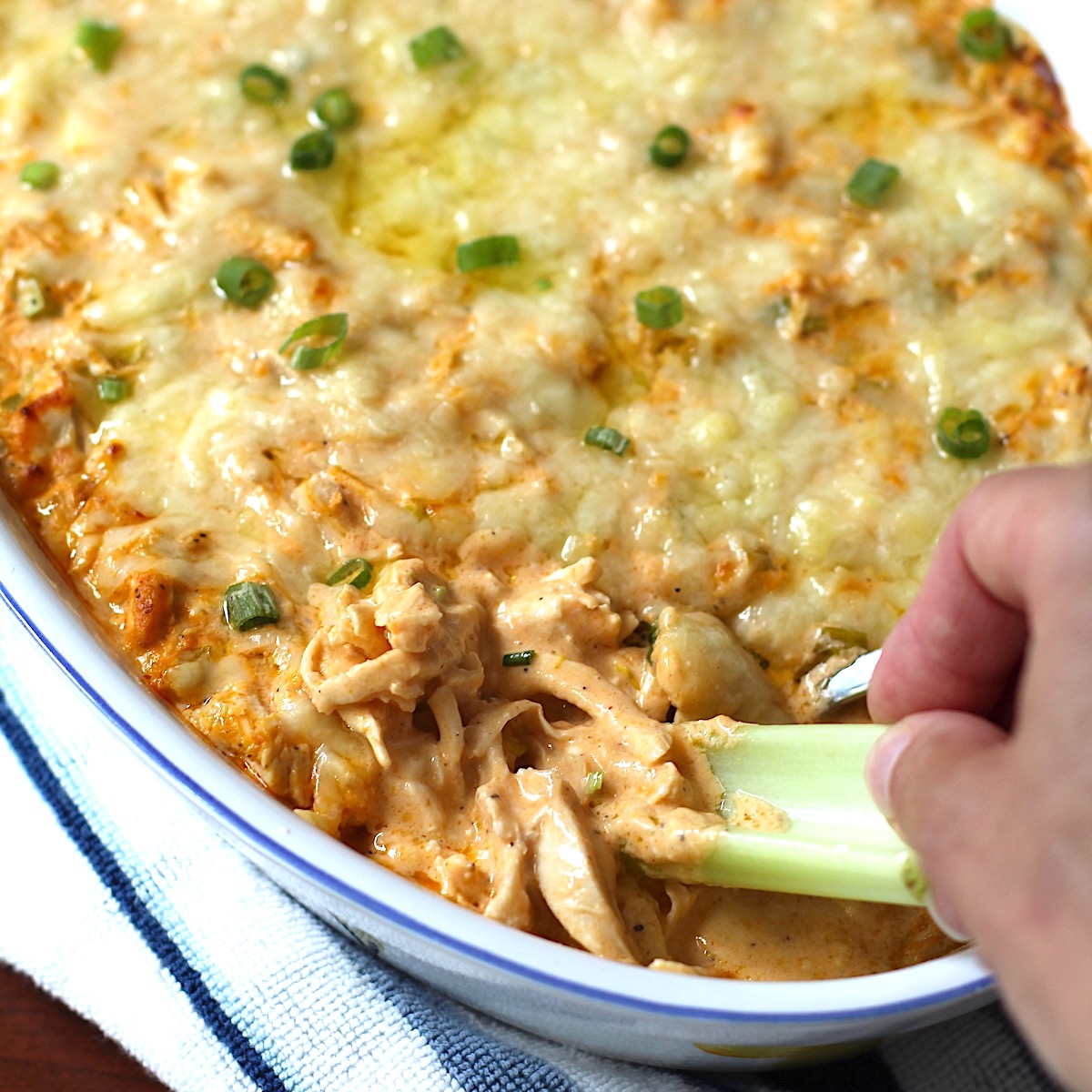 Hand with celery scooping Buffalo Chicken Dip with greek yogurt in serving dish with spoon in it.
