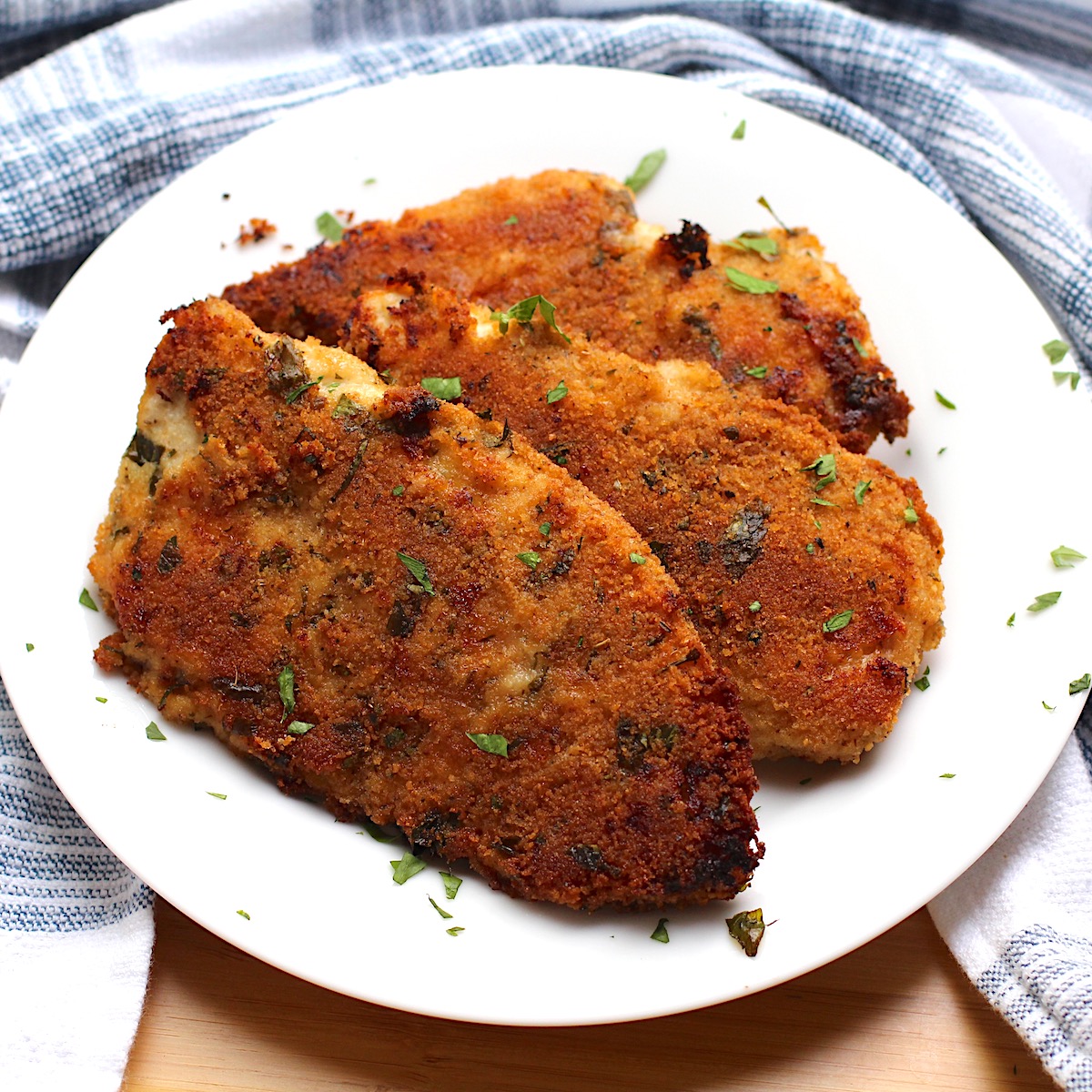 Three golden brown, baked Italian Chicken Cutlets fanned out on a plate with parsley sprinkled on top. Plate is surrounded by a blue and white towel.