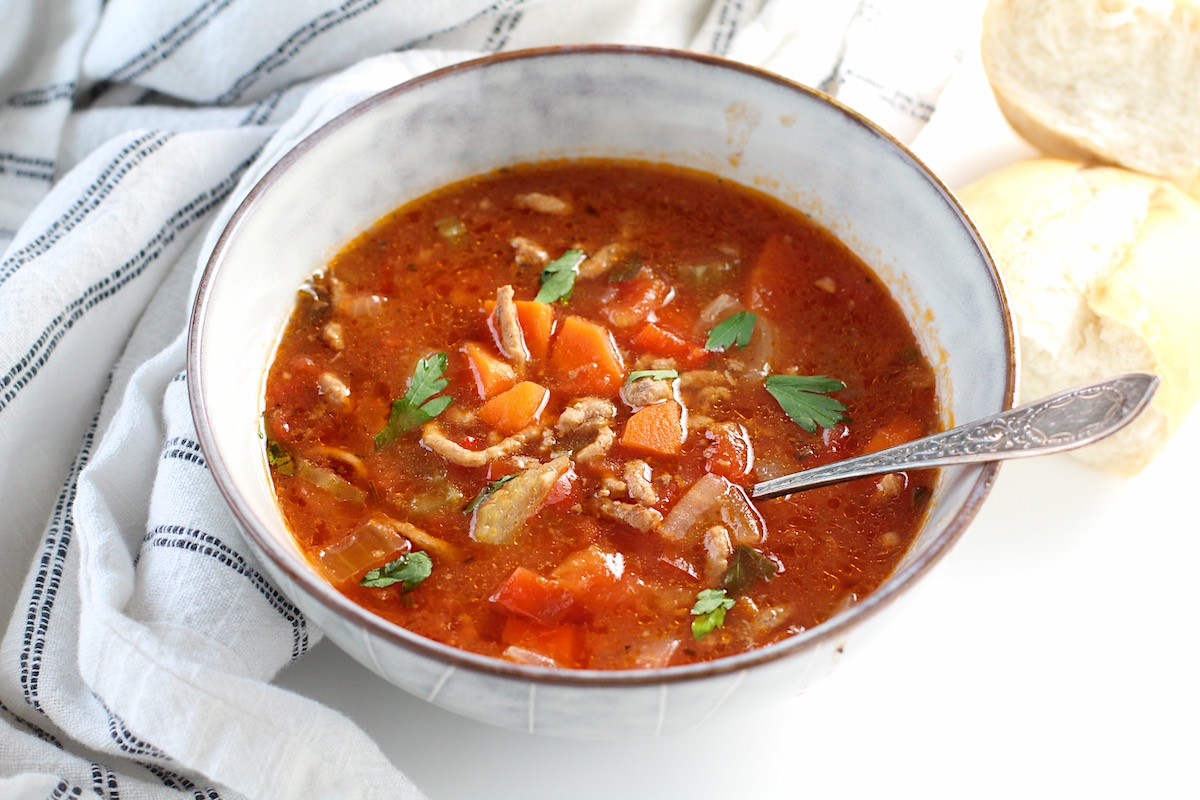 Ground Beef Vegetable Soup Recipe in a bowl with spoon on counter with blue and white towel around. It's a perfect fall meal that's comforting, hearty, and delicious with loads of veggies! Make it in the slow cooker or on the stove.