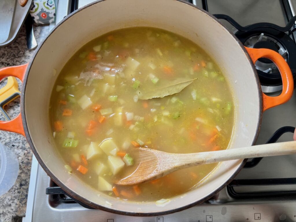 Diced potatoes, carrots, celery, onion, broth, and bay leaf in a pot cooking with wooden spoon for Chicken Potato Soup Recipe.