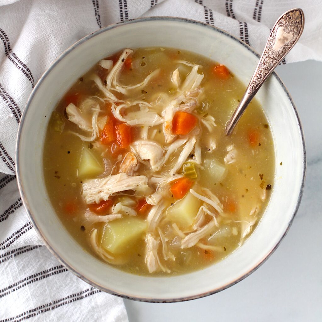 Creamy Chicken Potato Soup Recipe with carrots and celery in a bowl with a spoon on counter with towel. It's comforting, hearty, and delicious.  It's a great dinner for busy families!