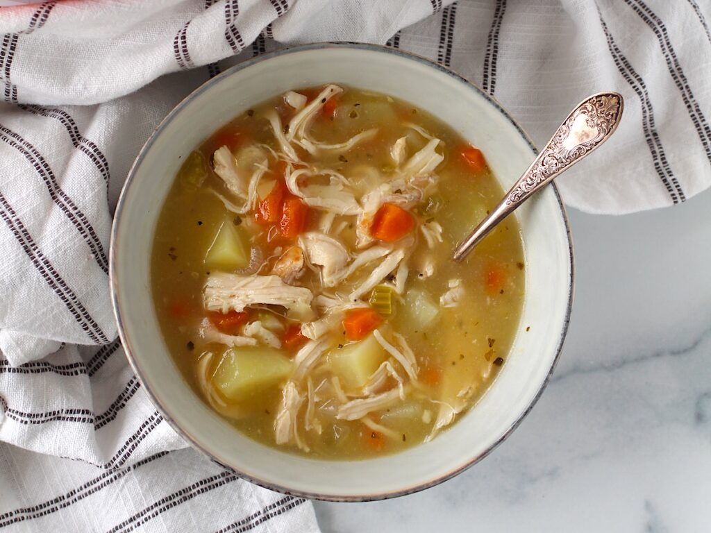 Creamy Chicken Potato Soup Recipe with carrots and celery in a bowl with a spoon on counter with towel. It's comforting, hearty, and delicious.  It's a great dinner for busy families!
