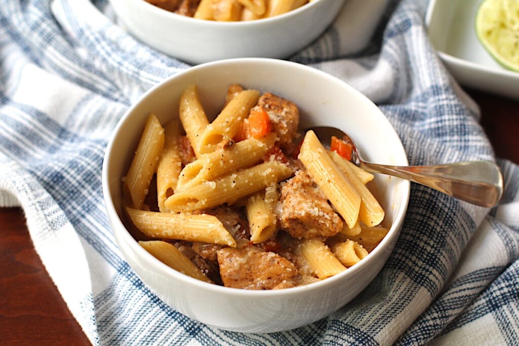 Two bowls of Chipotle Chicken Pasta with penne pasta, diced red pepper, parmesan cheese sprinkled on top, and a fork in the bowl on top of kitchen towel.