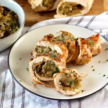 Stuffed chicken sliced on a plate with another stuffed chicken breast cut in half in the background, and a bowl of stuffing to the side. Stuffing stuffed chicken breast recipes are easy and delicious!