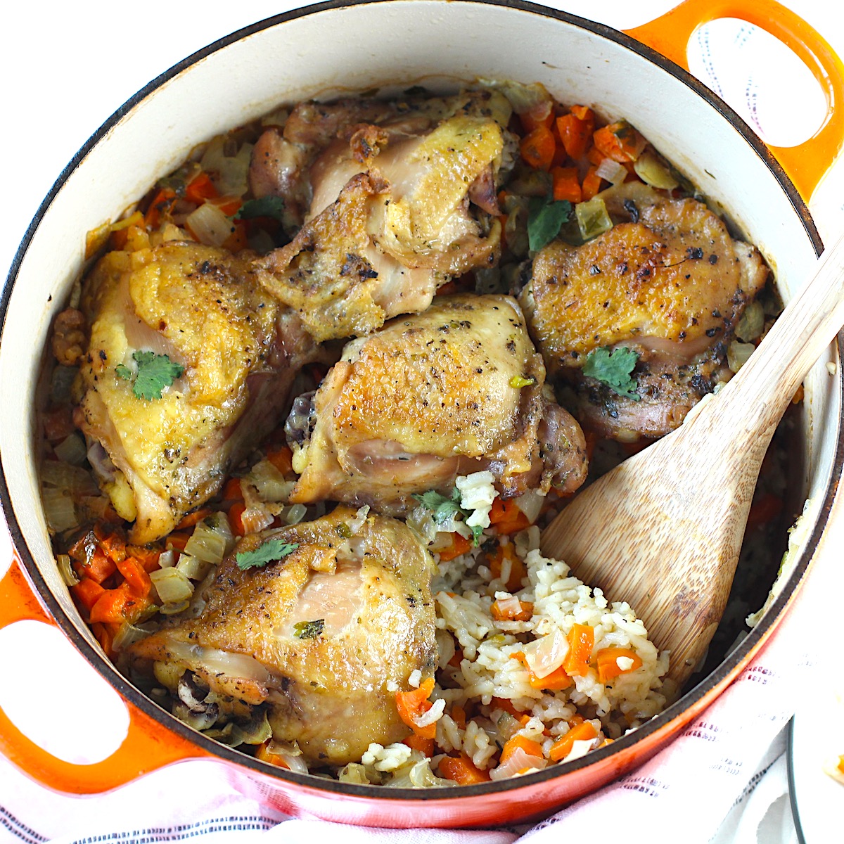 Roasted Chicken Rice in a pot with 5 crispy chicken thighs on top of rice with carrots and onions and a wood spatula scooping the rice. This recipe is an easy and delicious family dinner that you can make on any busy weeknight or cook entirely ahead!
