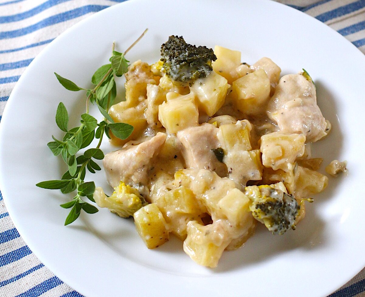 Chicken Broccoli Potato Casserole on a plate with a sprig of fresh oregano and the casserole dish in the background, all on a blue and white striped tablecloth.