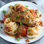 Smothered Chicken with Bacon, and Tomatoes and fettuccine on a plate with pan in background. The chicken is smothered in a thickened sauce infused with smokey and salty bacon flavor.  Cherry tomatoes give a sweet and tangy pop.