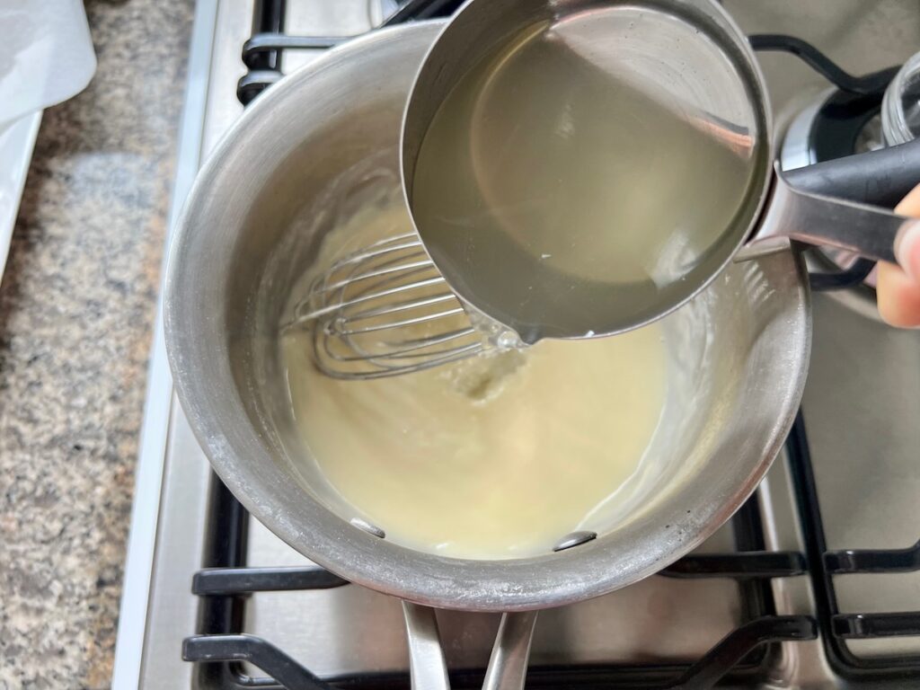 Broth being added to melted butter and flour combined in a saucepan with whisk mixing them.