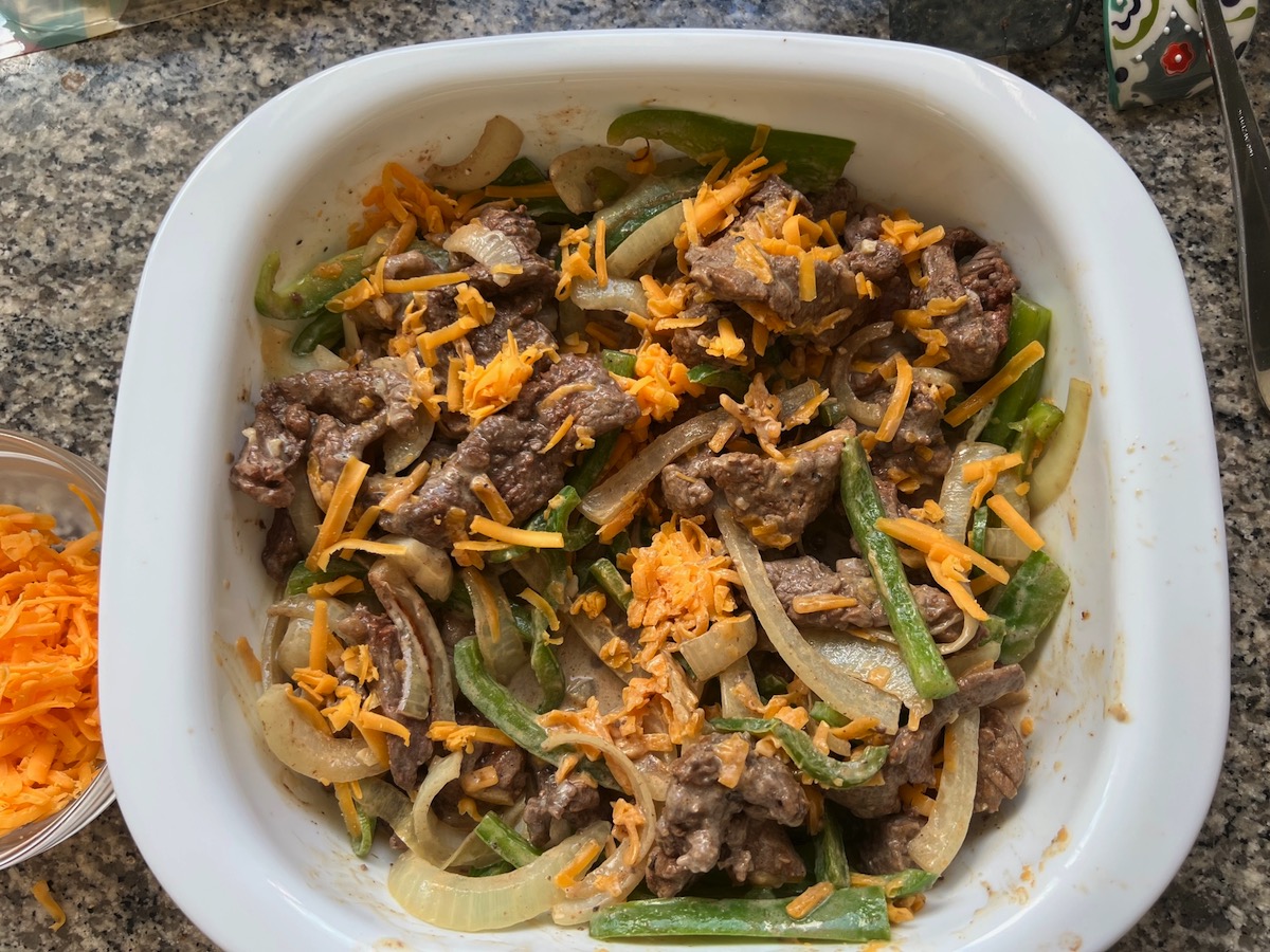 Cream cheese sauce and shredded cheddar mixed with flank steak, onions, and peppers in casserole dish for Philly Cheesesteak casserole recipe with Steak.