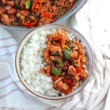 Thai Basil Pork Recipe with mini red pepper slices in a bowl over rice on the counter with a towel with skillet in background.
