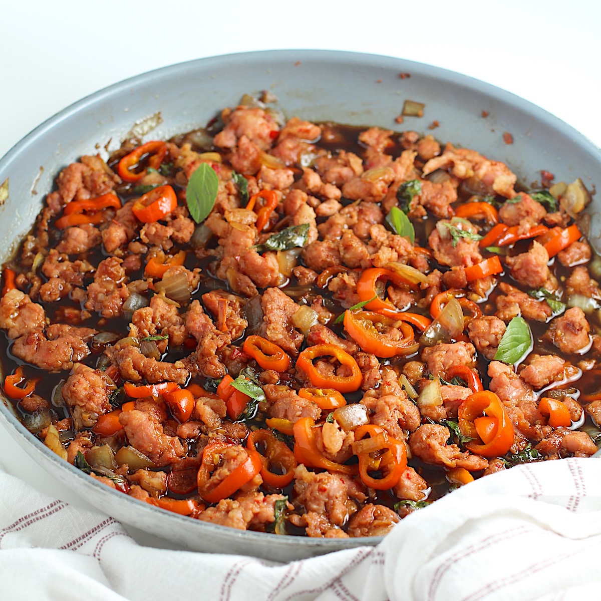 Thai Basil Pork Recipe with mini red pepper slices in a skillet on the counter with a towel wrapped around the handle.