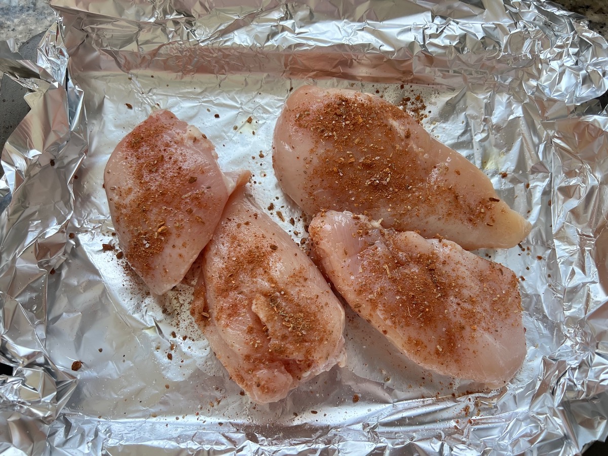 Raw seasoned skinless chicken breast on aluminum lined sheet pan to be roasted for the for crockpot white chicken enchilada casserole.