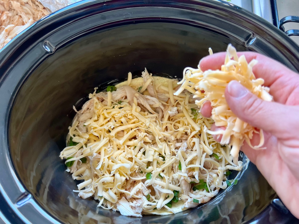 Hand adding shredded cheese on top of shredded chicken that is layered on corn tortillas for crockpot white chicken enchilada casserole.