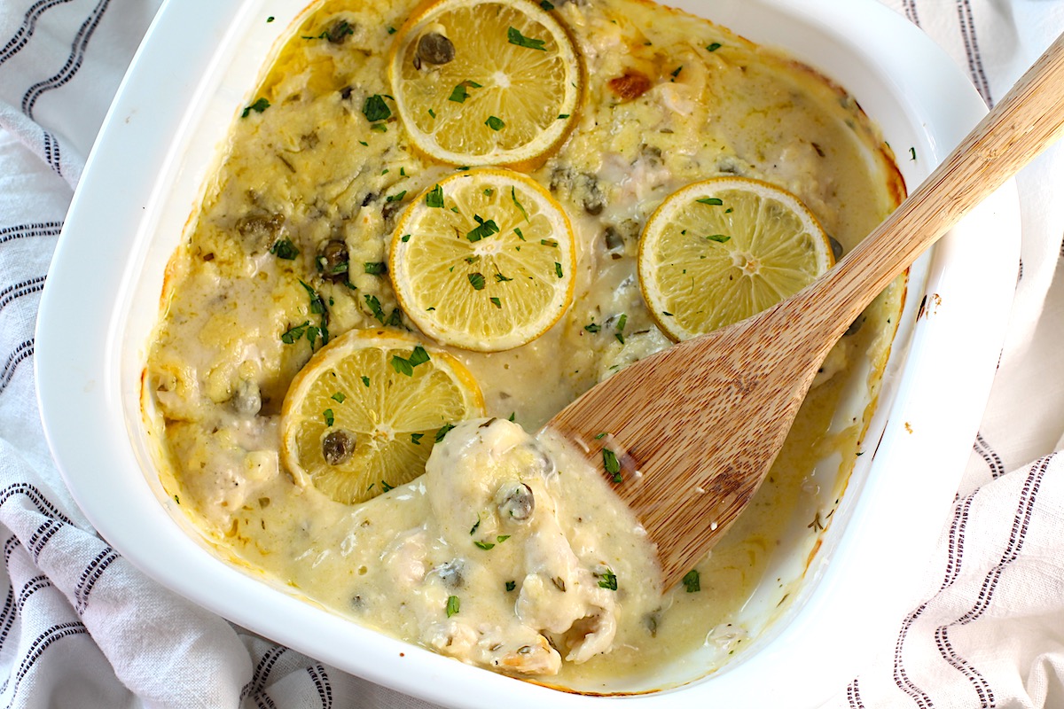 Creamy Chicken Piccata Casserole Recipe in a casserole dish with lemon slices and parsley on top. A wooden spatula is scooping a piece of chicken in the creamy sauce up from the corner.