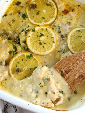 Creamy Chicken Piccata Casserole Recipe in a casserole dish with lemon slices and parsley on top. A wooden spatula is scooping a piece of chicken in the creamy sauce up from the corner.