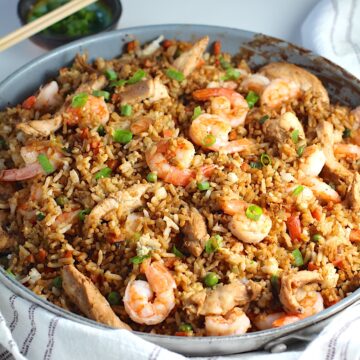 Shrimp and Chicken Fried Rice in a large frying pan on a table with a blue and white towel. Sliced scallions are sprinkled on top and in a small bowl behine the pan with chopsticks on top.