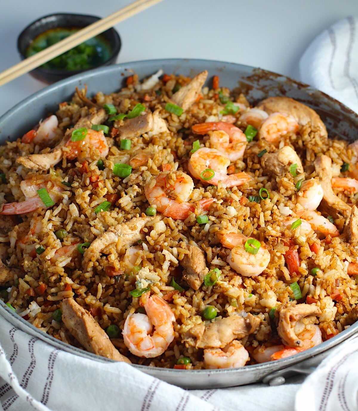 Shrimp and Chicken Fried Rice in a large frying pan on a table with a blue and white towel.  Sliced scallions are sprinkled on top and in a small bowl behine the pan with chopsticks on top.