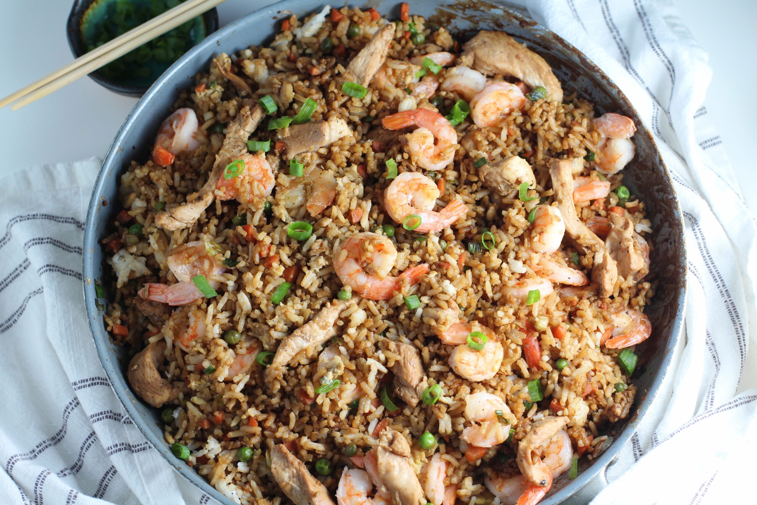 Shrimp and Chicken Fried Rice in a large frying pan on a table with a blue and white towel. Sliced scallions are sprinkled on top and in a small bowl behine the pan with chopsticks on top.