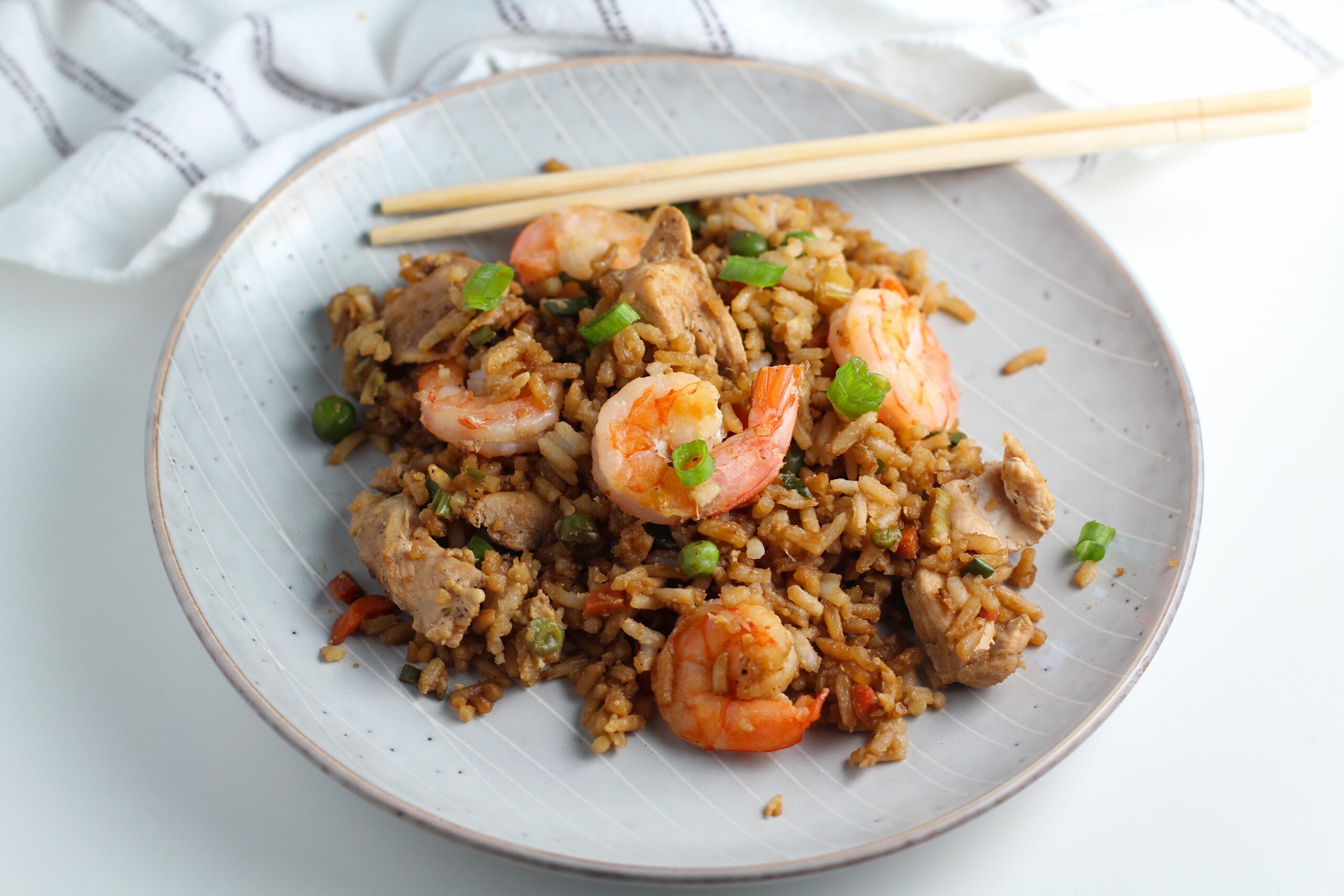 Shrimp and Chicken Fried Rice on a plate with chopsticks on a table with a blue and white towel. Sliced scallions are sprinkled on top.