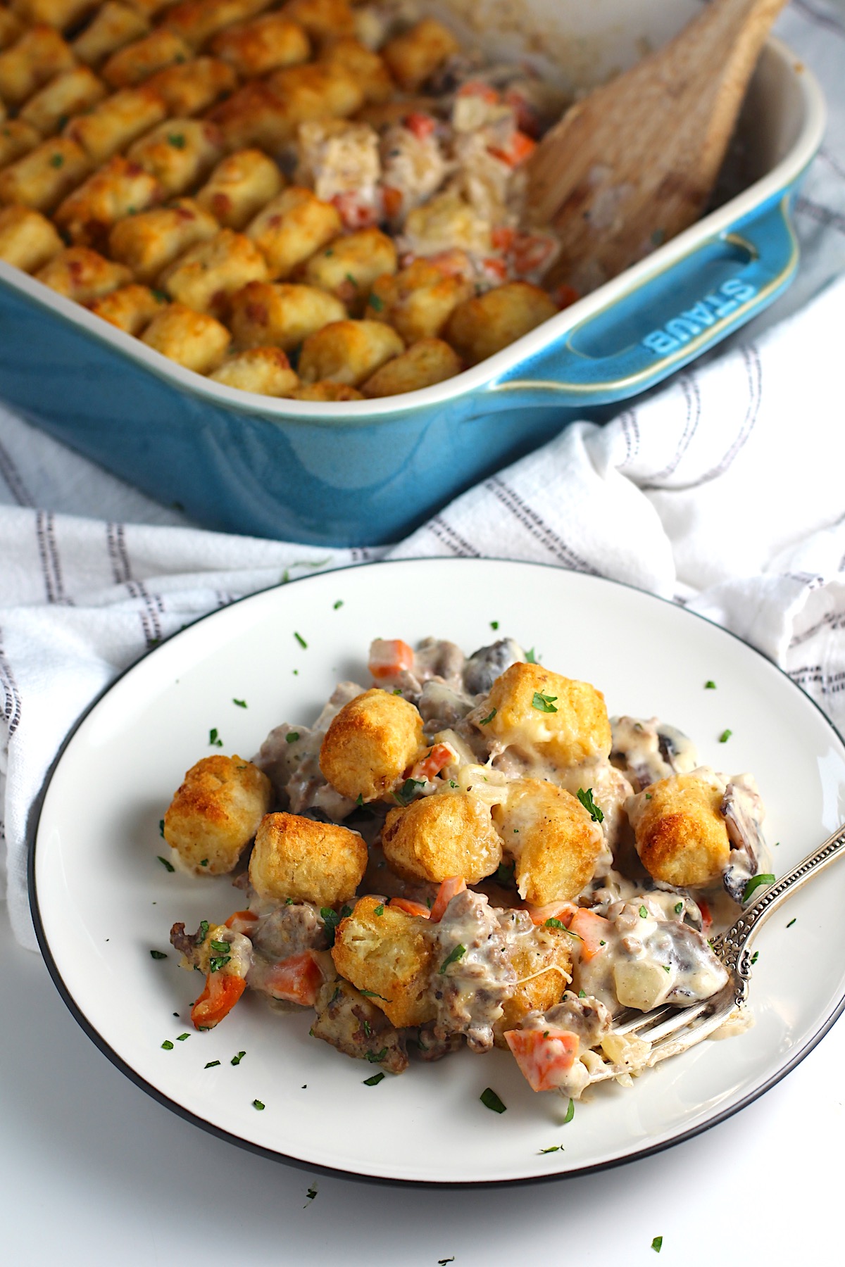 Creamy and cheesy hamburger tater tot casserole recipe on a plate with a fork and parsley sprinkled on top. The casserole dish is in the background.