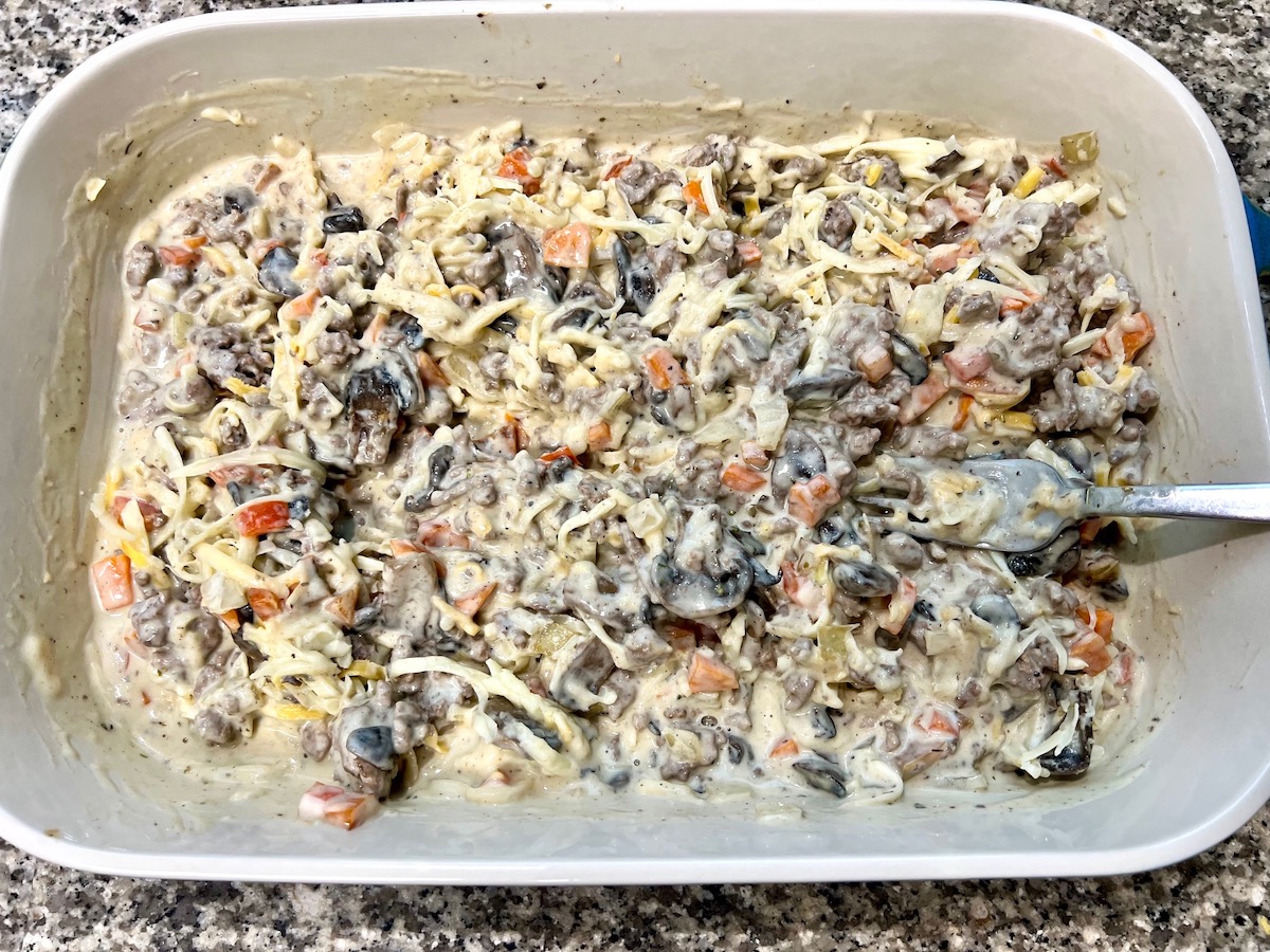 Thickened sauce and cheese mixed into mushroom slices, diced red pepper, diced carrots, diced onion, and ground beef mixed and cooking in bottom of casserole dish, during the preparation of the Cheesy Hamburger Tater Tot Casserole Recipe.