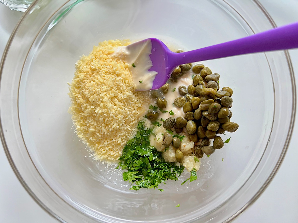 Mayonnaise, parmesan, parsley, capers, lemon juice, and garlic, in a clear mixing bowl with spatula for the Creamy Chicken Piccata Casserole recipe.