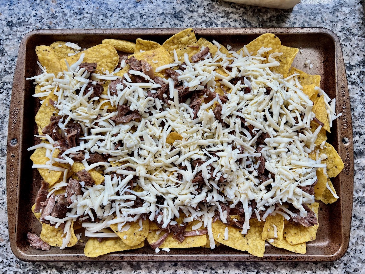 Shredded white cheddar on top of shredded Beef layered on top of yellow corn tortilla chips on a large sheet pan on counter for Birria Nachos.