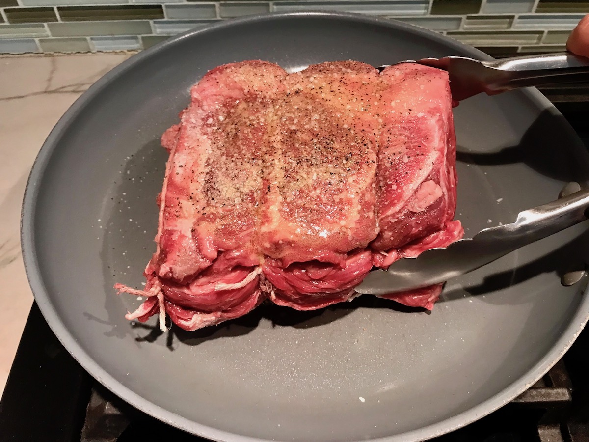 Seasoned beef chuck roast being placed onto a hot frying pan with tongs to be seared before slow cooking for Birria Nachos.