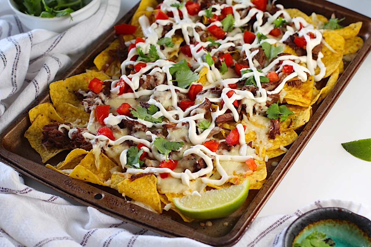 Birria Nachos on a sheet pan with yellow corn toritlla chips topped with tender shredded beef, melty cheese, a drizzle sour cream, fresh diced tomatoes, cilantro leaves, and a lime wedge in the corner of the pan.