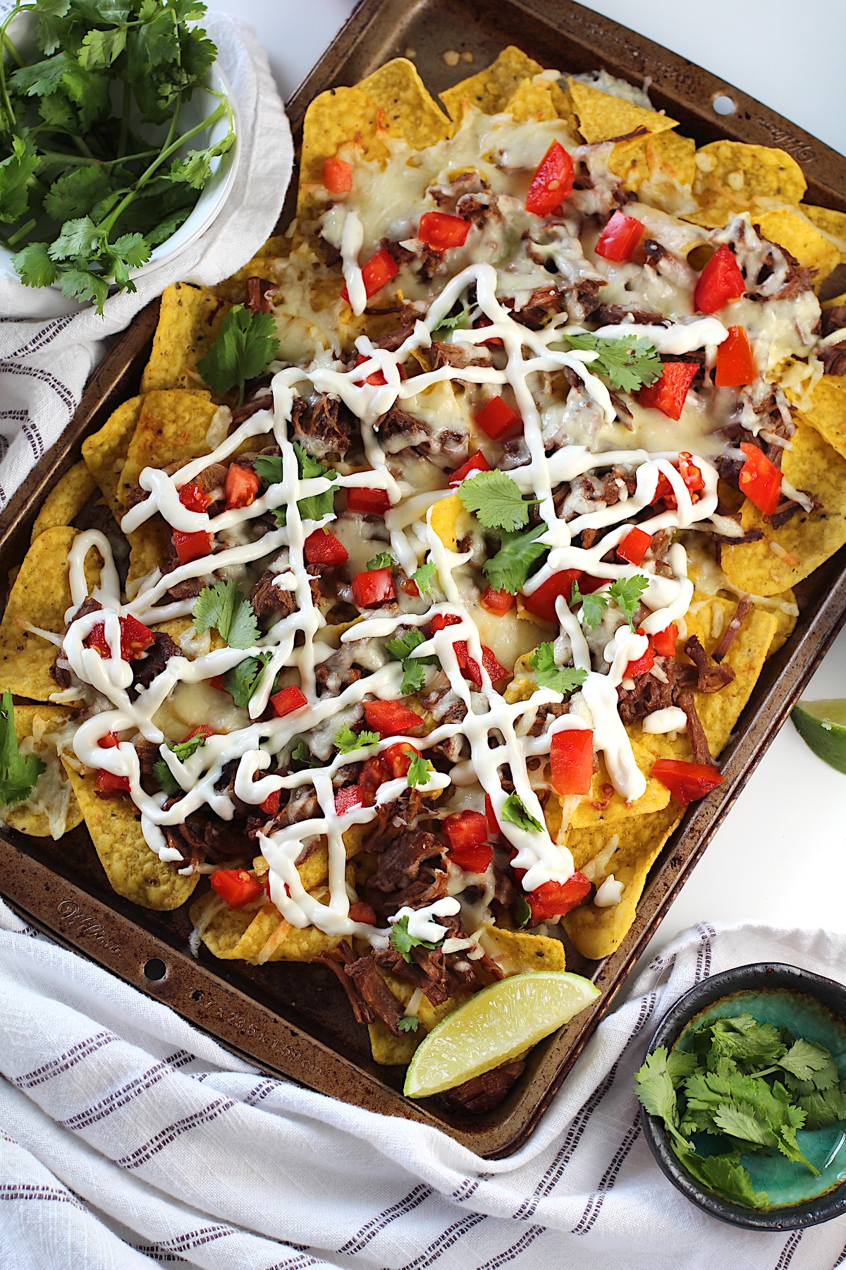 Birria Nachos on a sheet pan with yellow corn toritlla chips topped with tender shredded beef, melty cheese, a drizzle sour cream, fresh diced tomatoes, cilantro leaves, and a lime wedge in the corner of the pan.