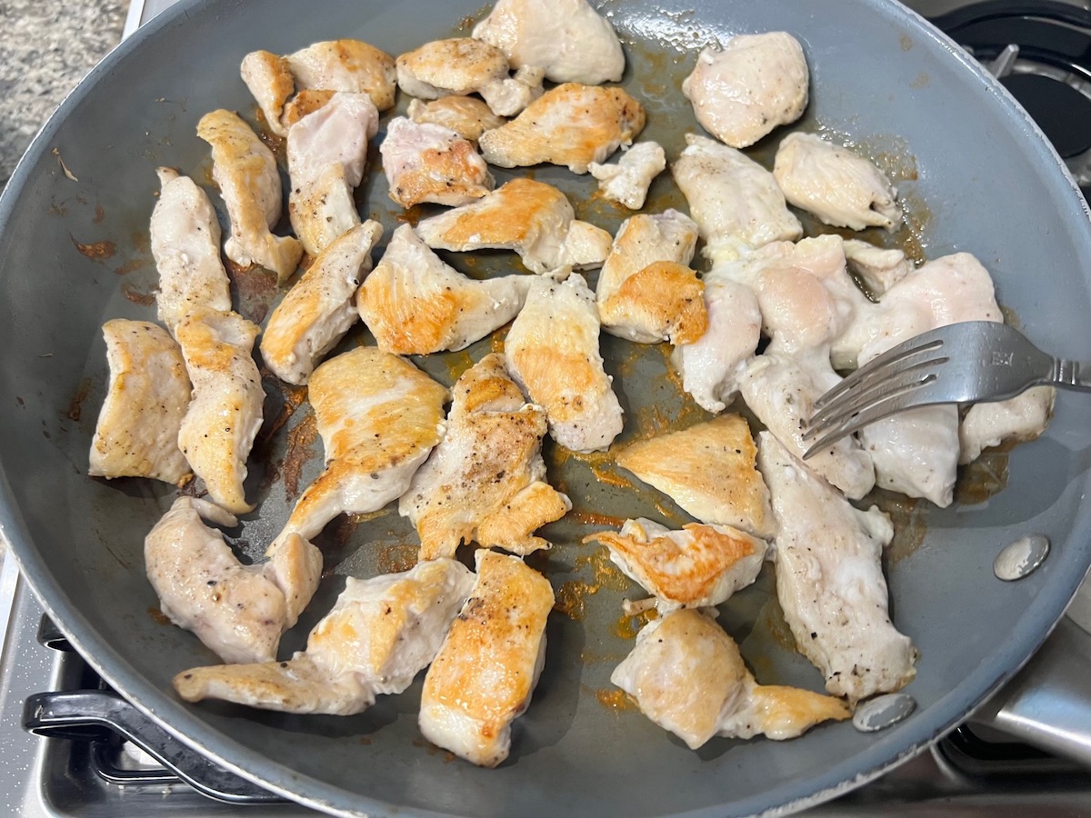 Chicken breast pieces being browned in a large frying pan with a fork flipping over the pieces for Chicken and Vodka Pasta recipe.