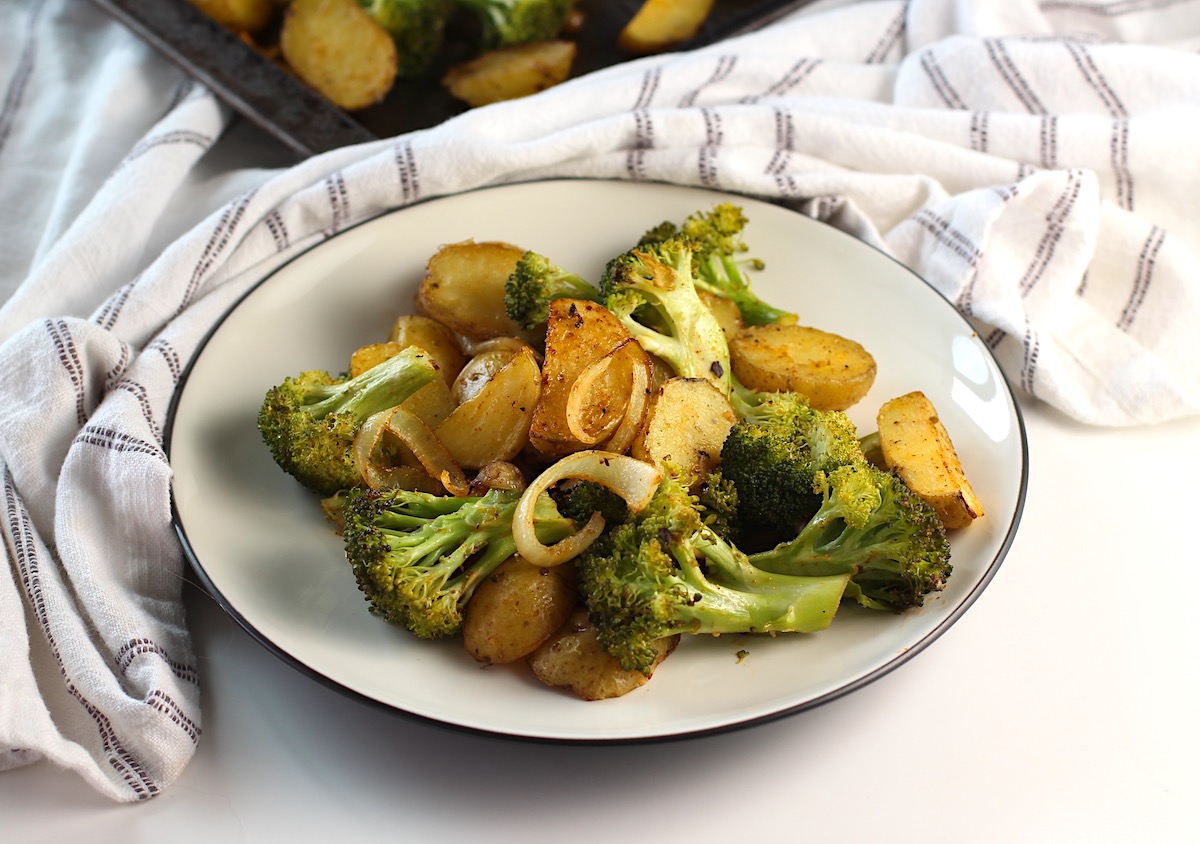 Plate of crispy, golden brown, roasted potatoes, broccoli, and onions with a sheet pan of more in the background.