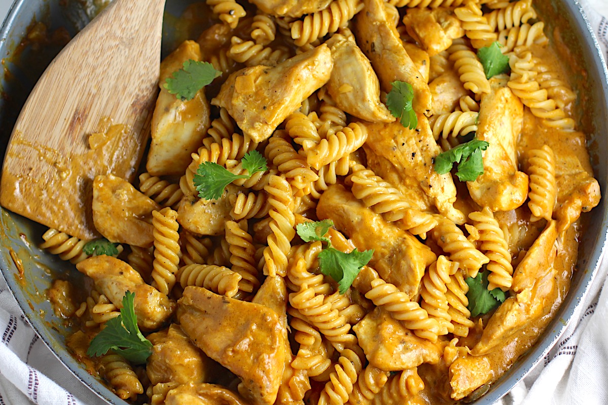 Skillet of butter chicken pasta topped with chopped cilantro and a wooden serving spoon resting in the skillet.