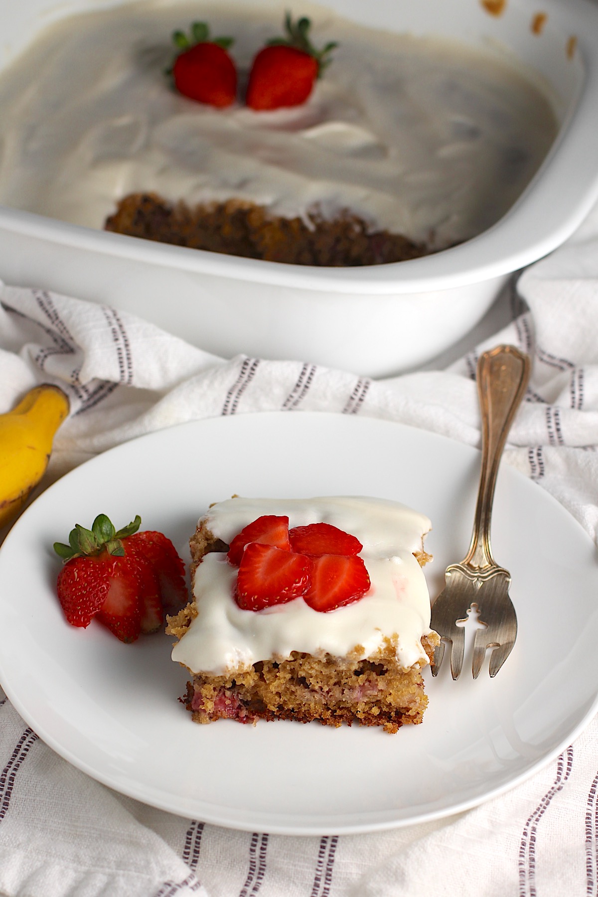 Piece of Strawberry Banana Cake with Vanilla Icing on a plate with a fork with sliced strawberries on top and a strawberry sliced and fanned out to the left.