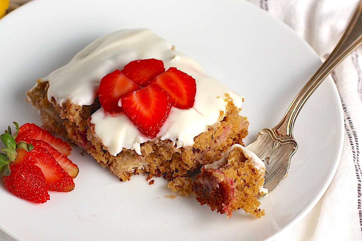 Piece of Strawberry Banana Cake with Vanilla Icing on a plate with a fork holding a bite and sliced strawberries on top and a strawberry sliced and fanned out to the left.