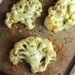 Four Cheesy Cauliflower steaks roasted on a sheet pan with cheese melted on top and edges browned and crispy.