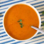 Sweet Potato and Red Pepper Soup in two bowls with fresh oregano leaves around the table and garnishing the soup in one bowl. Blue and white striped tablecloth in background.