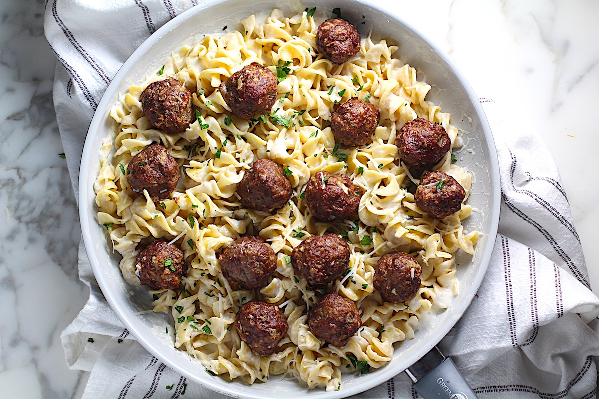 Alfredo and meatballs with meatballs nestled into creamy sauce and noodles in a frying pan on towel on counter.