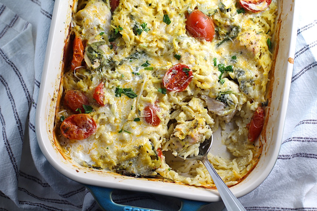 Cheesy Chicken Orzo Bake with grape tomatoes, spinach, and a creamy sauce in a casserole dish with spoon scooping some out at the corner.