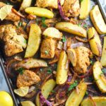 Crispy, golden brown Greek Lemon Chicken and Potatoes with red onions and fresh parsley garnish on a sheet pan on the counter. Whole lemon sitting to the left on the counter.
