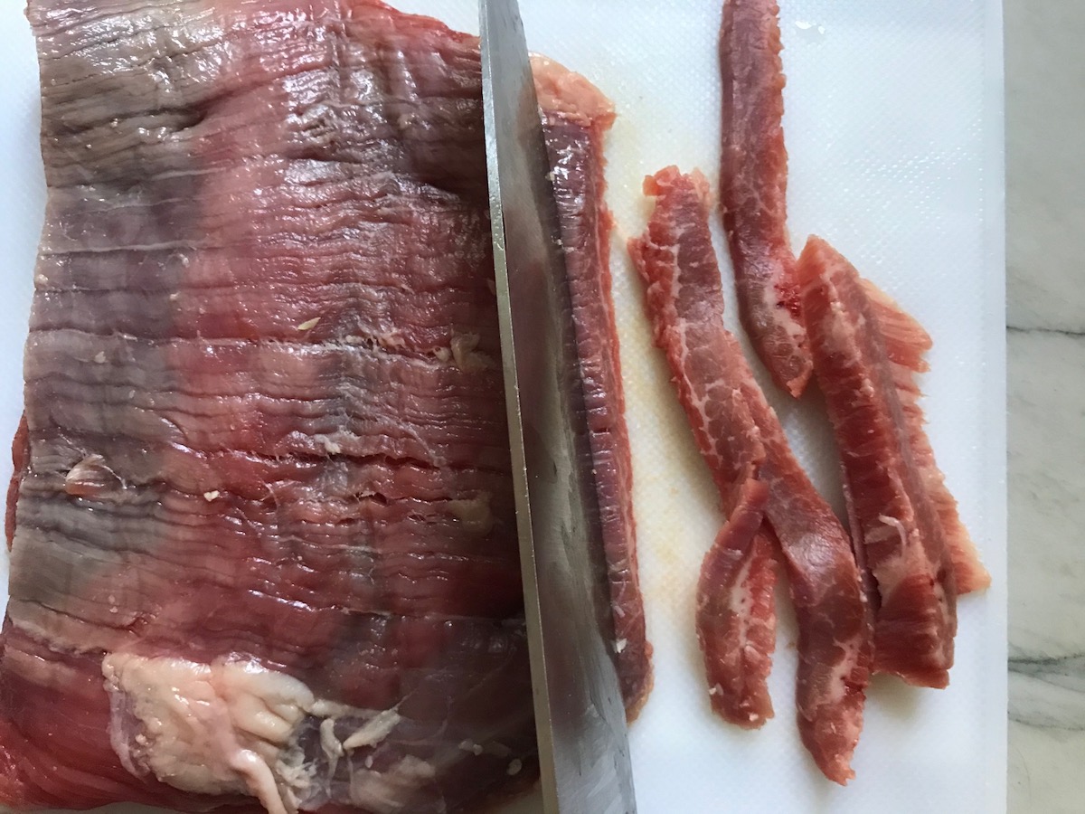 Chef's knife slicing raw flank steak against the grain into thin strips on cutting board for beef and broccoli noodles recipe.