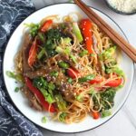 Steak, broccoli, red pepper, scallions, and noodles stir fried and on a plate with sesame seeds on top and chopsticks resting on the side for Beef and Broccoli Noodles recipe.