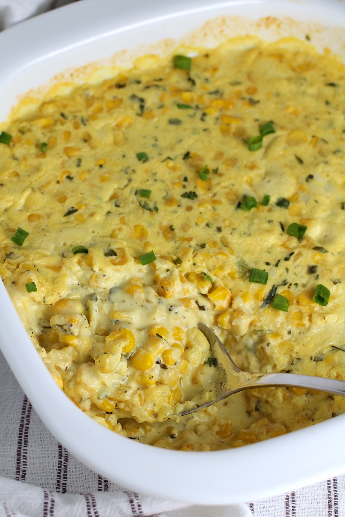 Creamy Mexican Street Corn Casserole with sliced scallions sprinkled on top in a dish on blue and white striped towel with serving spoon scooping from the corner of the casserole.