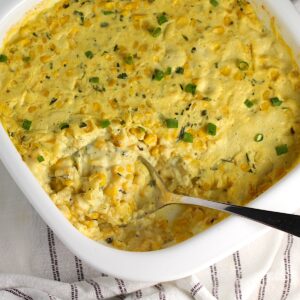 Creamy Mexican Street Corn Casserole with sliced scallions sprinkled on top in a dish on blue and white striped towel with serving spoon scooping from the corner of the casserole.